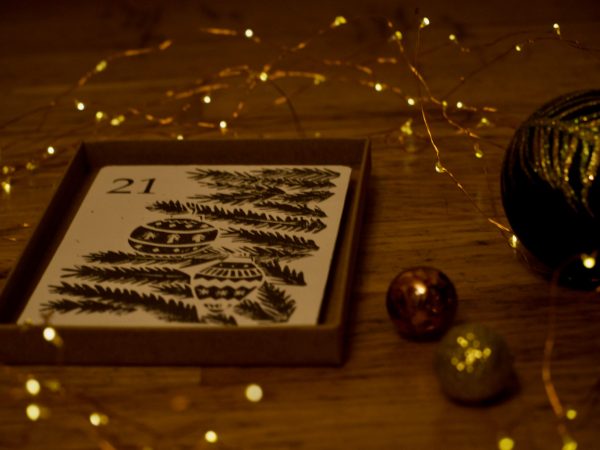 A card displaying a christmas tree and the number 21 sits in a cardboard box. To the right are fairy lights and baubles.